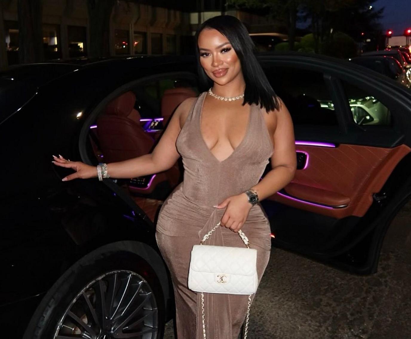 A woman in a light brown dress stands in front of a car, with a shell necklace and straight black hair.