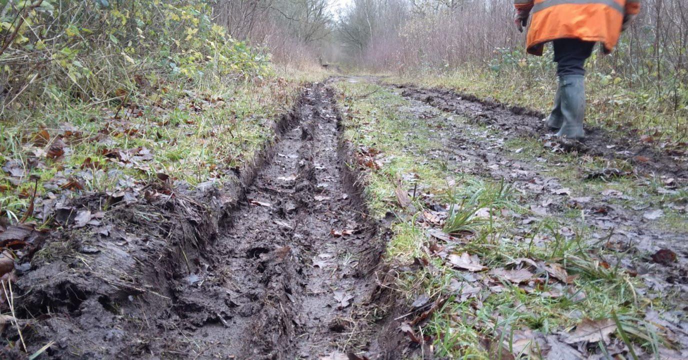 Close up on a deep tyre track in a pathway through a woodland, with a person in wellies and orange high vis walking nearby