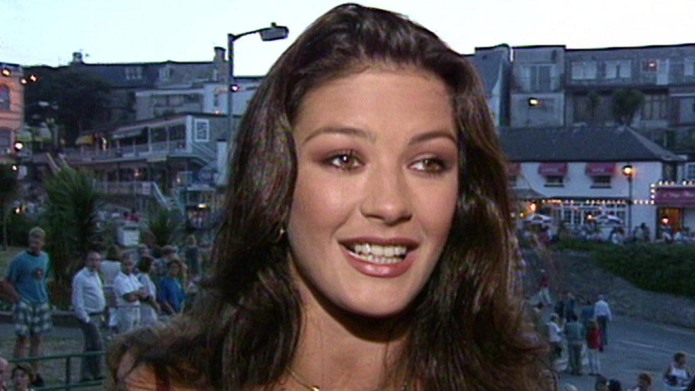 Catherine Zeta-Jones speaking to the camera with people standing around in the background at twilight.