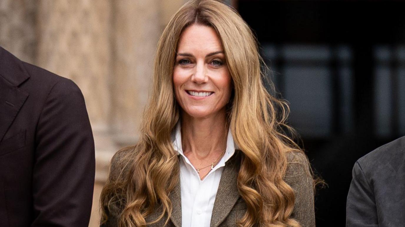 Kate, who has long curly dark blonde hair, seen smiling at cameras 
