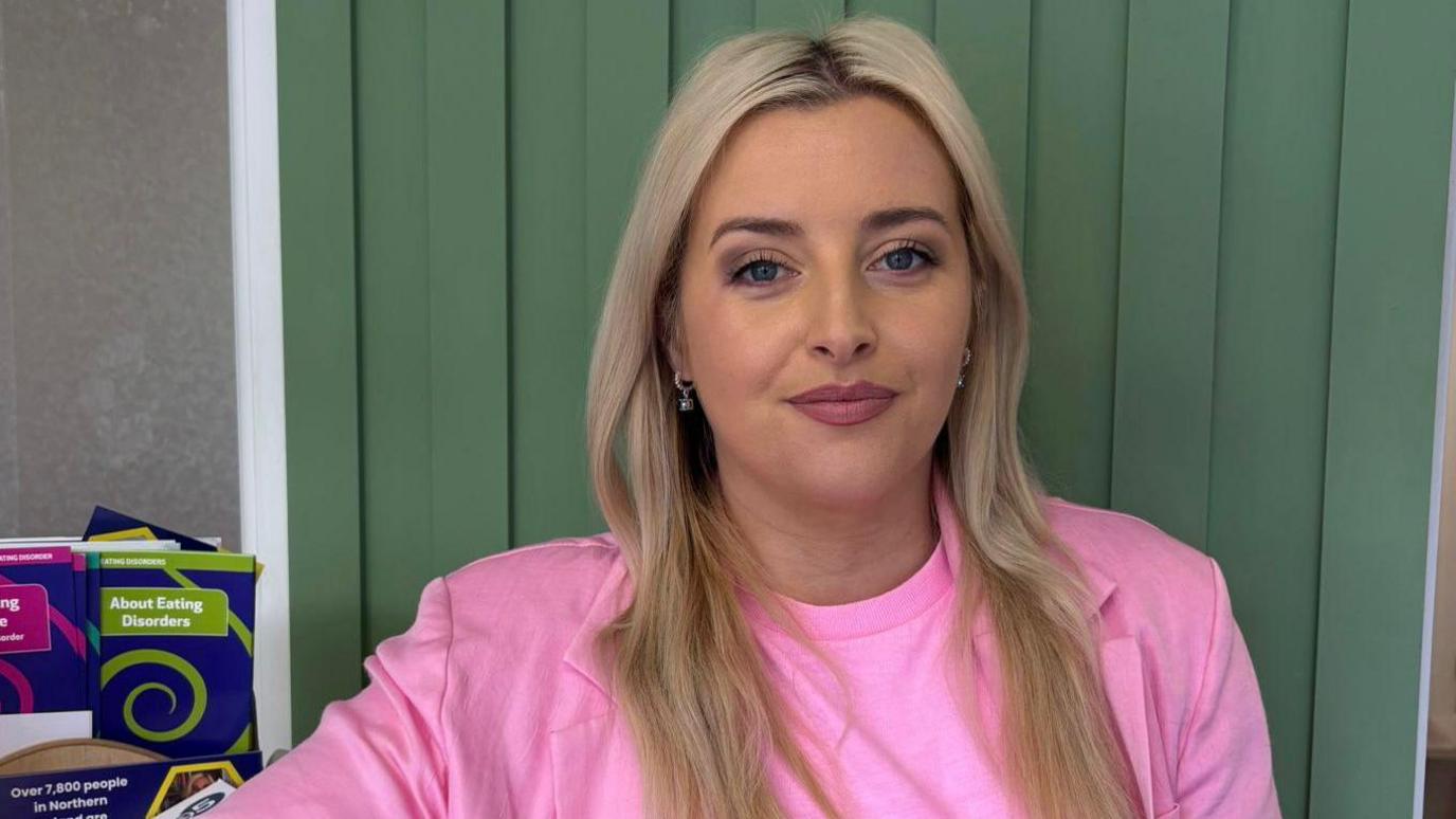Órlaithí Flynn has blonde hair, a side parting and gold earrings. She is wearing a 
bright pink t shirt and a pink jacket and is standing in front of a wood-panelled wall which is painted in green