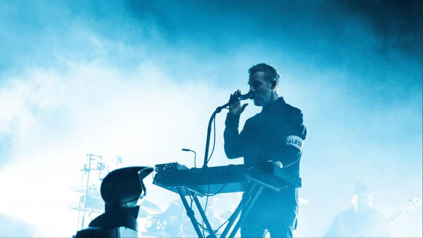 Massive Attack's 3D stands at a keyboard and speaks into a microphone on stage at the ACT 1.5 show, his band stands behind him and he is surrounded by smoke