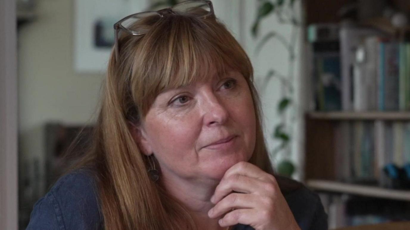 A woman with long strawberry-blonde hair and fringe looks thoughtfully away from the camera with her hand on her chin