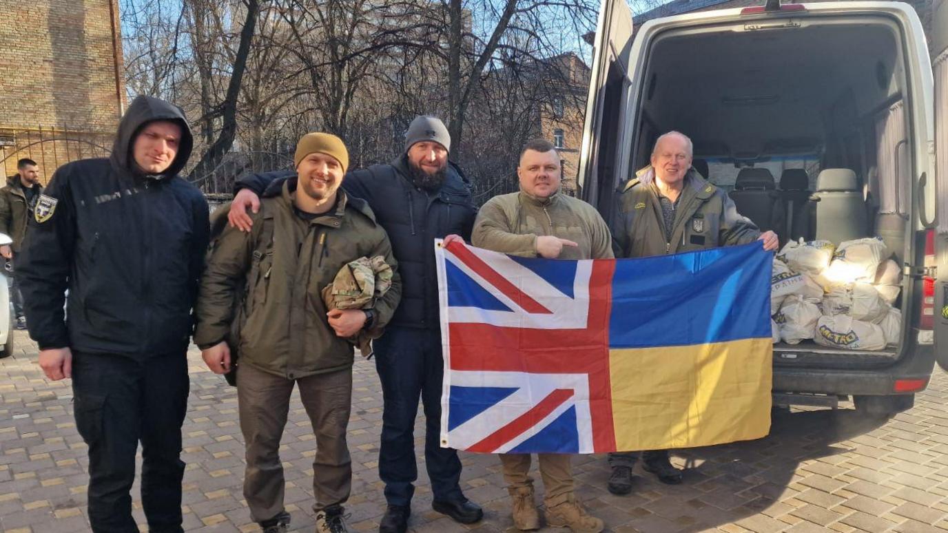 Five men in a line, with two holding a flag - half Union Flag and half Ukrainian flag - next to a van with bags of supplies.