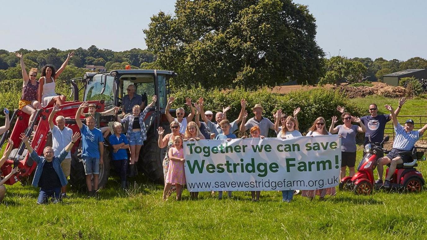 Two dozen campaigners are pictured with arms aloft in a field on a sunny day in 2021. The group includes men, women and children. Some are sitting on a tractor and some are holding a large banner that reads: "Together we can save Westridge Farm!"