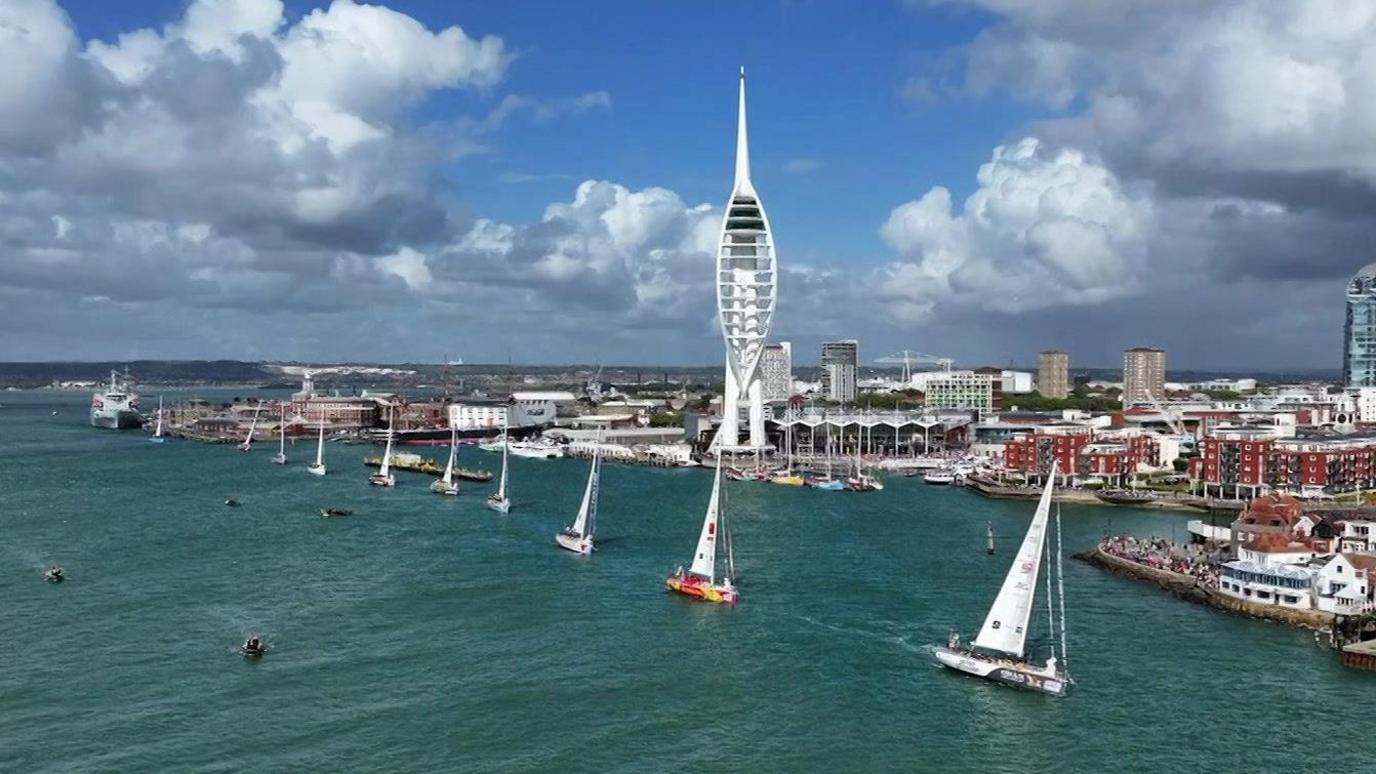Drone shot of the Clipper yachts leaving Portsmouth, filing past the Spinnaker tower - a white sail-shaped observation tower on the waterfront.