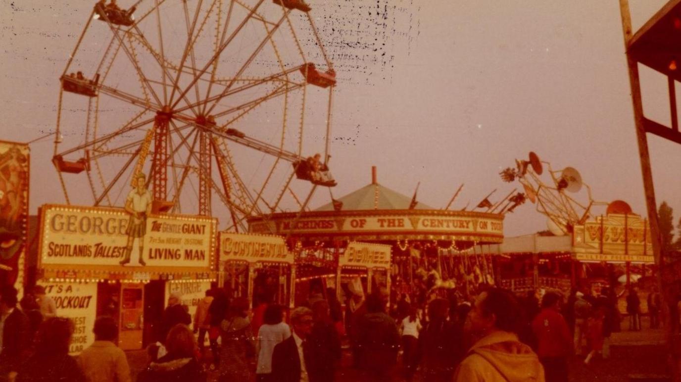 A picture with a red tint from the 1970s showing fairground rides and an attraction called Scotland's Tallest Man.