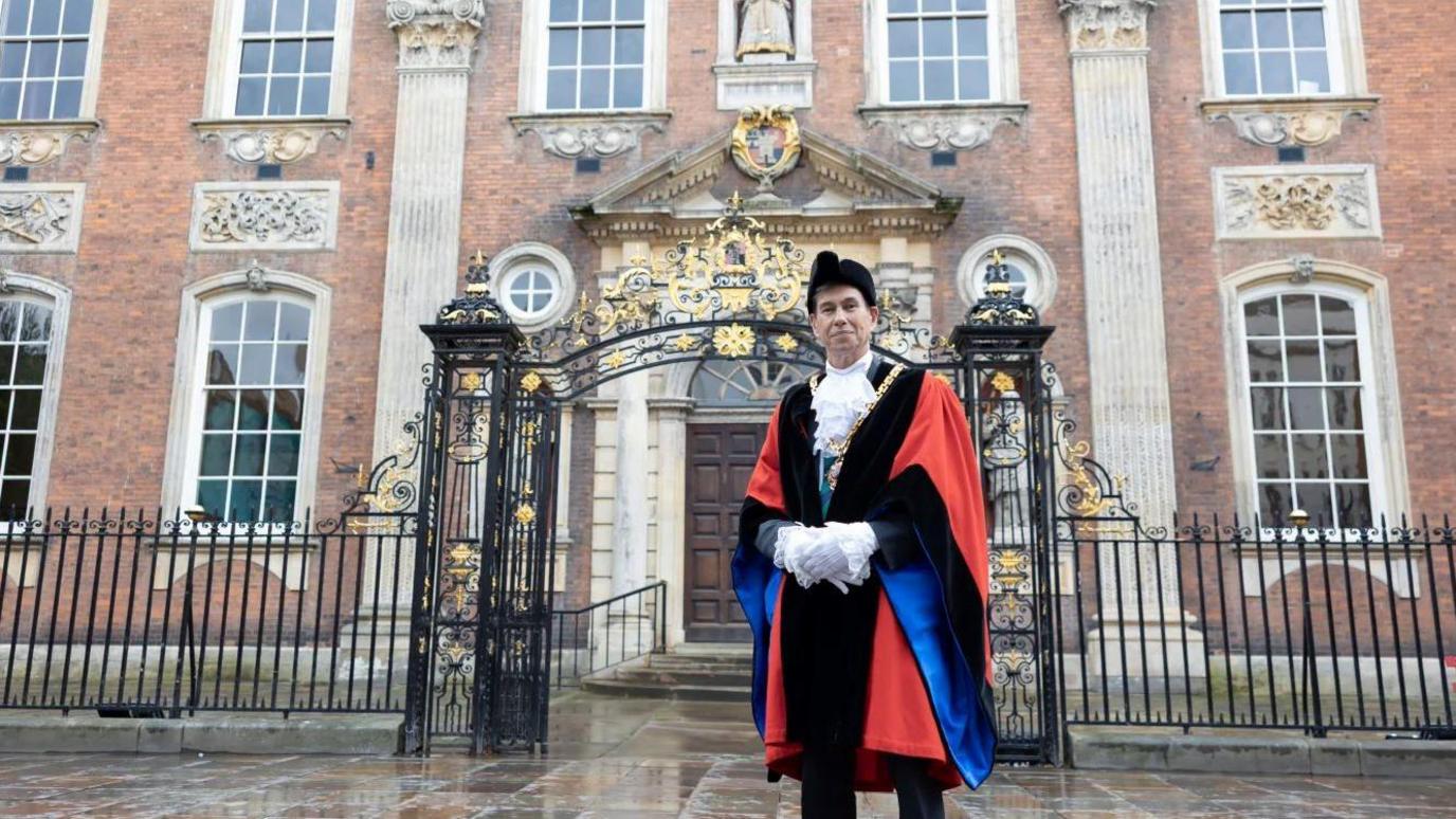 A man in official mayoral dress, which includes a black hat, gold chain and red, blue and black robes. Behind him is a brown and white building and black gates with gold decorations.