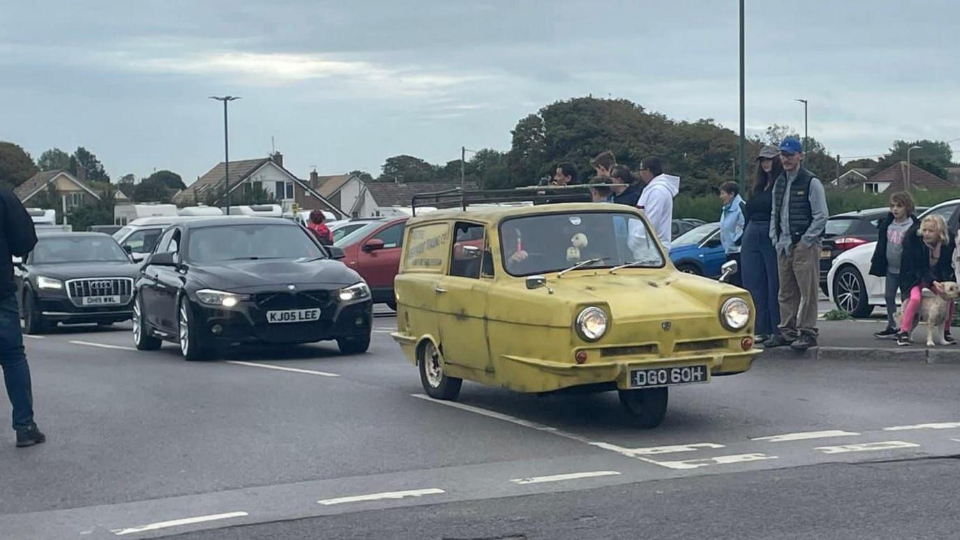 A yellow Morris Minor car, or similar, with three wheels arrives at a give way line at a junction in a wet car park, followed by a black BMW and a black Audi. People are gathering to look at the cars as they drive by.