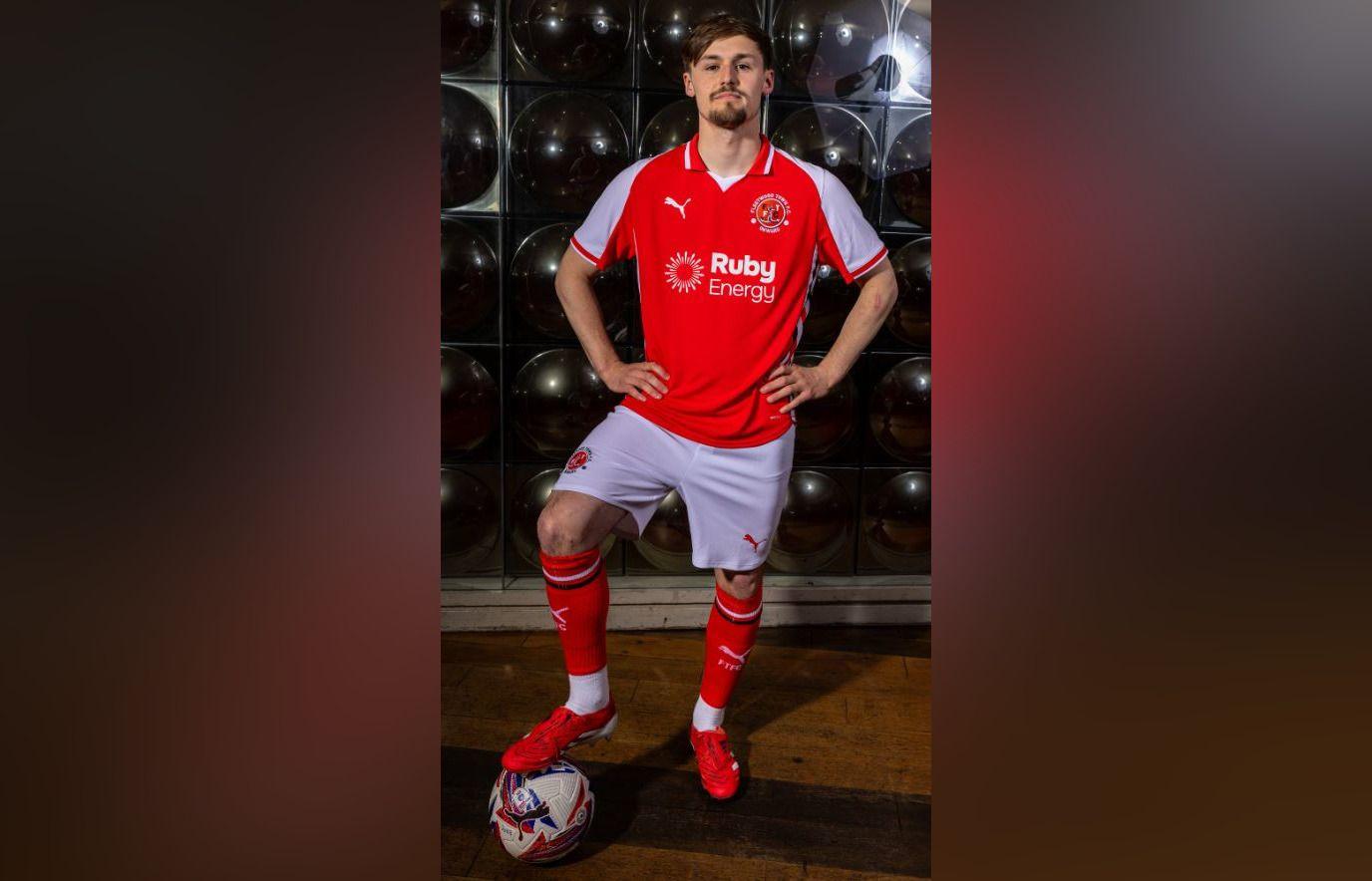 A man with short dark hair and a goatee beard stands with his hands on his hips and right foot resting on a football. He is wearing a red football shirt with white sleeves, across the front of which is a white Puma sign, the club crest and the sponsor Ruby Energy. He is also wearing white football shorts, red socks pulled up to his knees and red football boots.