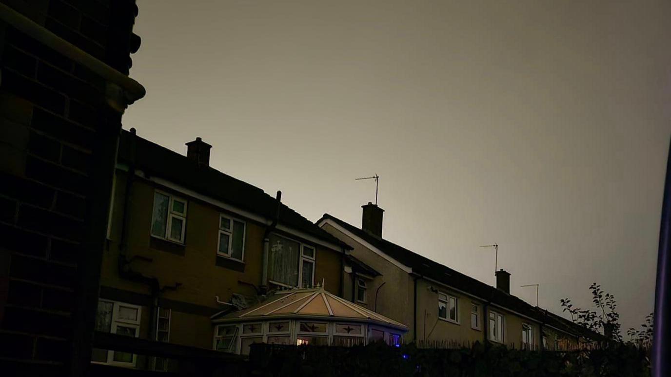 A glow in the night sky can be seen, in the foreground there are terraced houses