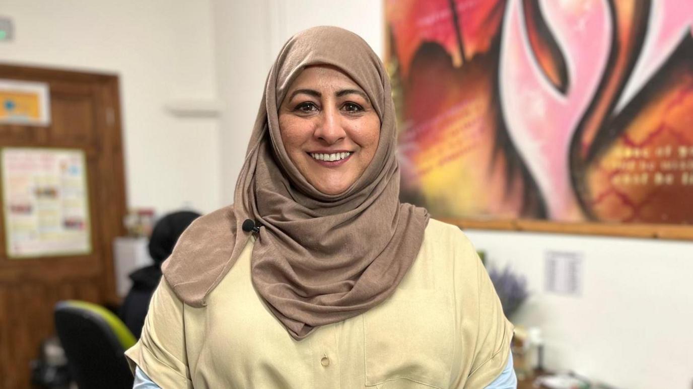 Mae Selima Bahadur yn sefyll mewn swyddfa gyda llun lliwgar ar y wal tu nol iddi. Mae'n gwisgo penwisg frown golau a chrys melyn.