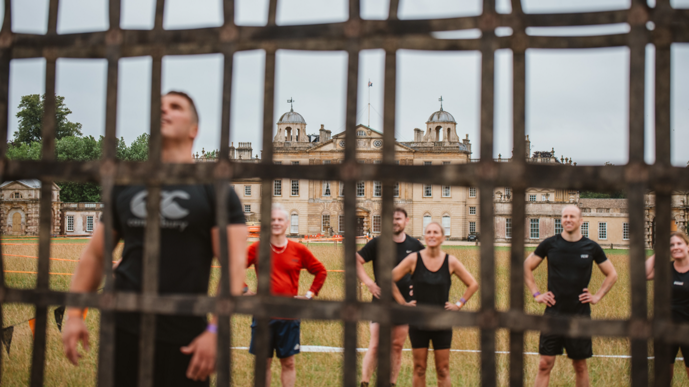 Participants lined up in black t-shirts and leggings or shorts on a lawn behind a steel-latticed structure, ready to scale it. Badminton House is in the background