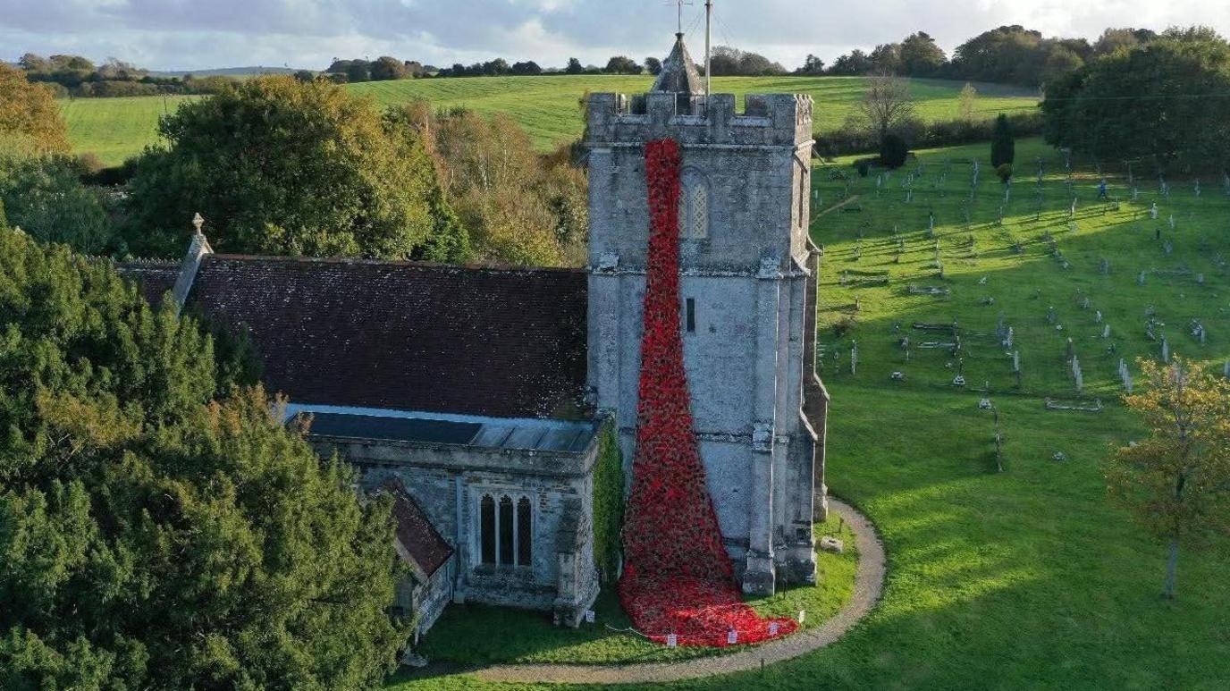 An aerial view of a medieval stone church in a green church yard with a cascade of red poppies hanging from the top of the bell tower