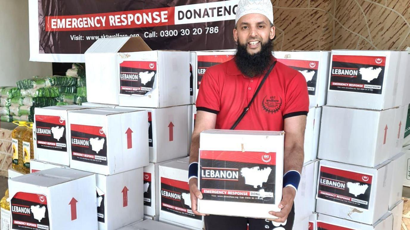 Nazim Ali in a red t-shirt holding a box of donations which say 'Lebanon emergency response'