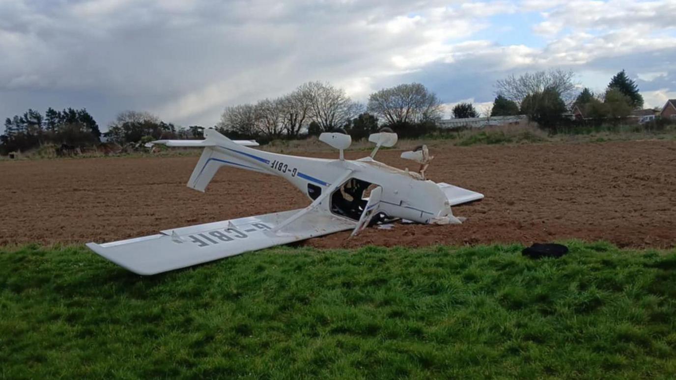 A white light aircraft which has landed on a patch of brown grass in an airfield.