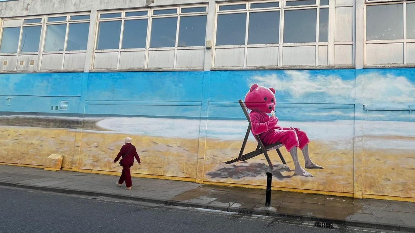 Mural of pink bear sitting on a deck chair on a golden beach with woman in red suit walking past