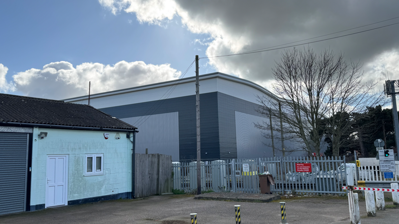 The corner of the warehouse can be seen next to a smaller building and just behind a railway