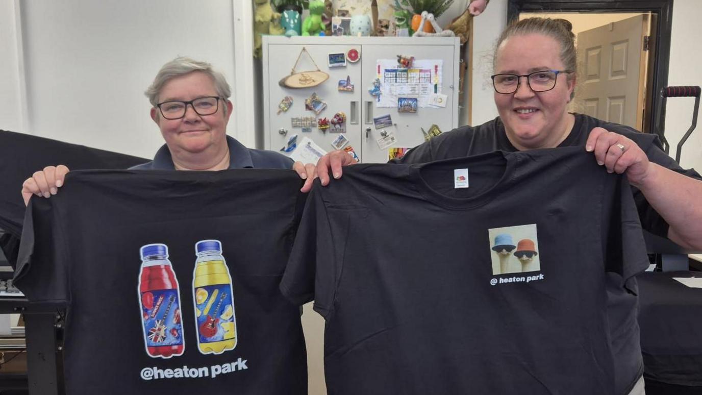 Two women hold up t-shirts they have printed. One has drinks bottles with rock guitar branding and the other shows figures in bucket hats and sunglasses. Both shirts feature Heaton Park branding.