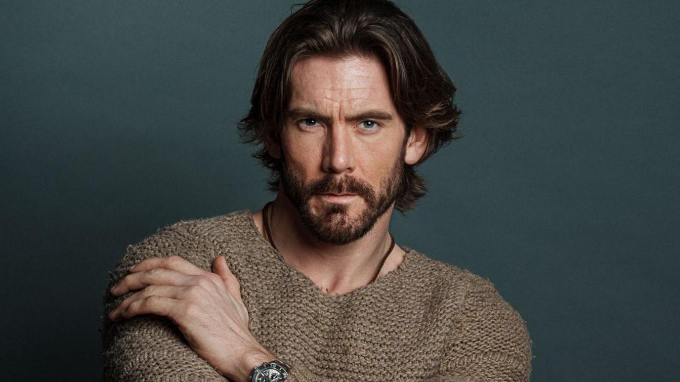 A headshot of James Wilkinson in a brown knitted jumper. He is posing while sat down in front of a dark green background. He has long brown hair and a dark beard.