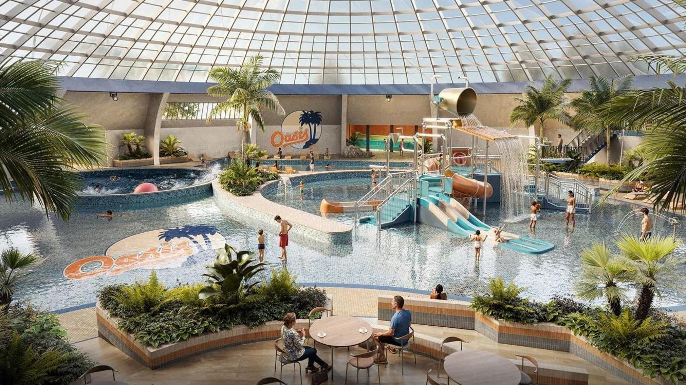 Image of the proposed inside of the refurbished Oasis Leisure Centre. It shows a pool with small water slides for young children and a large water bucket at height which creates a waterfall effect. There are palm trees and plants around the pool and also two sets of table and chairs in the foreground. The glass dome roof above the pool is visible.  