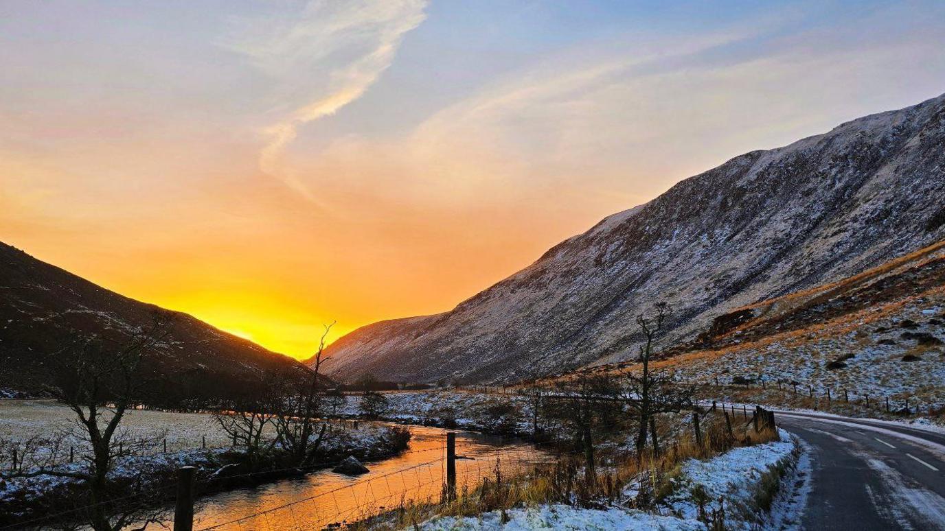 A slightly snowy road with a hill on one side and a river on the other. At the end is the sunrise which is yellow and orange.