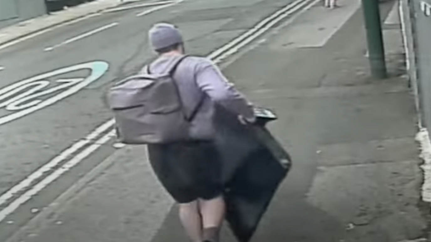 A CCTV image of a man carrying a large flat-screen TV. He is seen from the back and is wearing a grey jumper, hat and has a grey rucksack on his back. He is wearing black shorts.