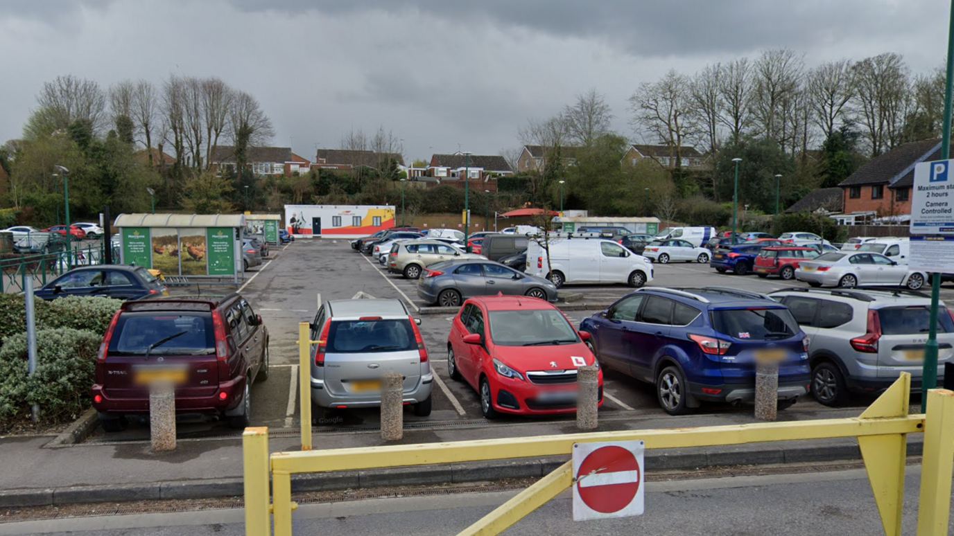 Morrisons car park in Warminster, with around 30 cars of various makes parked. There is a mobile unit at the back and some trolley parks. There is a no entry sign and barrier in the forefront of the picture.