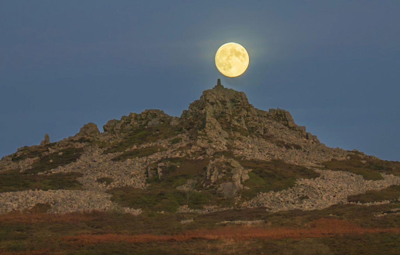 A rock formation on top of a hill with a pale yellow supermoon hovering over it.