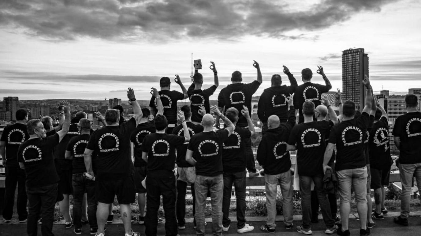 A group of men facing away from the camera making the 'OK' hand sign. They are all wearing Andy's Man Club t-shirts. The photo is in black and white. 