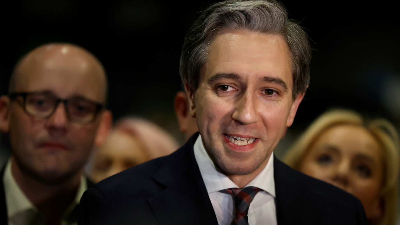 Simon Harris smiling with some people in the background. He is wearing a suit and tie