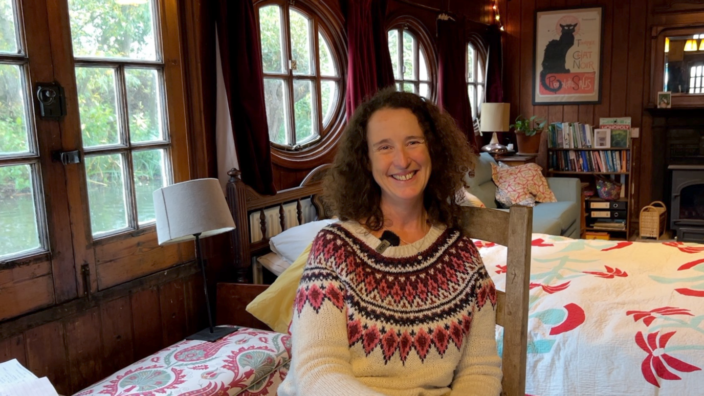 Zanna Hoskins smiling as she sits inside the Corpus Christi Barge. Behind her there is a bed, a sofa, a shelf with books and other pieces of furniture. 