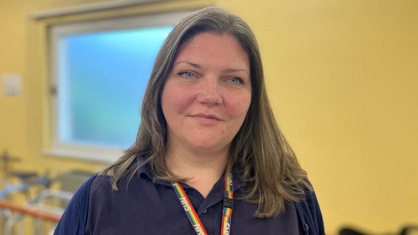 Specialist occupational therapist Vicky Carruthers smiles at the camera. She is wearing a rainbow lanyard. In the background rehabilitation equipment is blurred. The wall behind is yellow and there is a window.