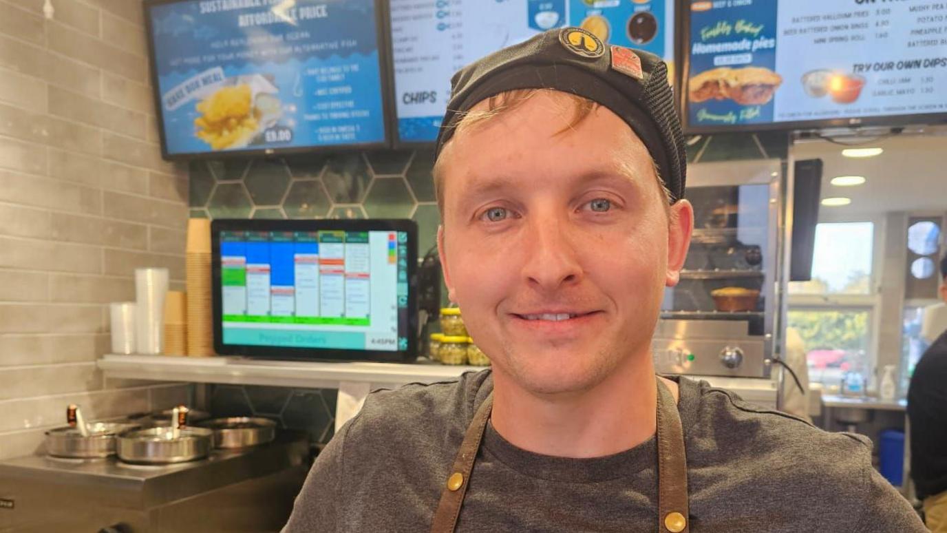 Steven Angell stands behind the chip counter. He has an apron on and a black hat. He's smiling at the camera. 