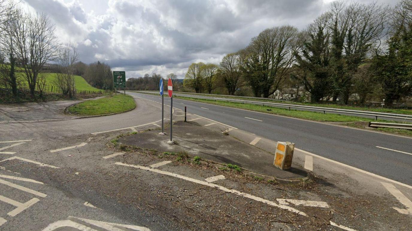 The A38 is empty with a slip road pictured. A no entry sign is seen facing toward the slip road to stop traffic entering the A38 in the wrong direction. There are also trees at the edge of the A38 running along the road.