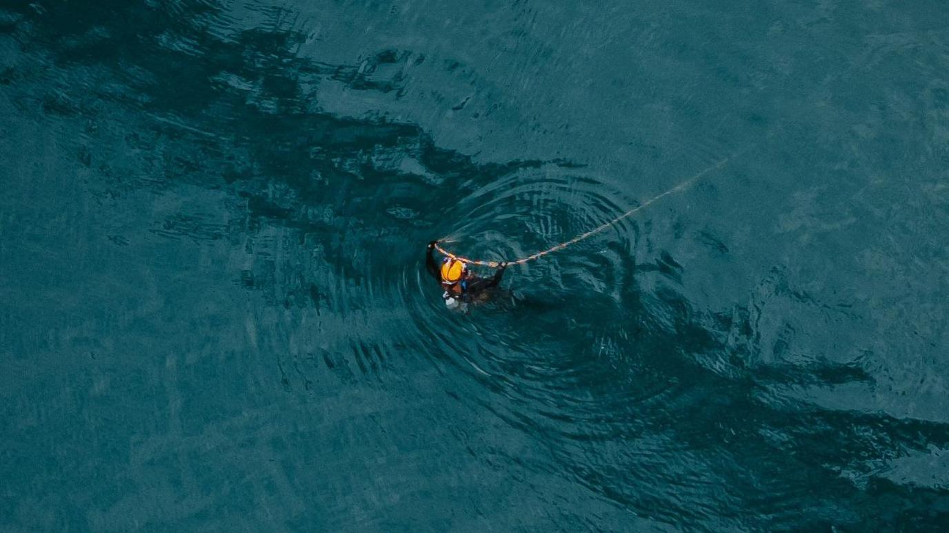 A diver or swimming is visible in the sea holding a rope or a wire. They have an orange hard hat on.