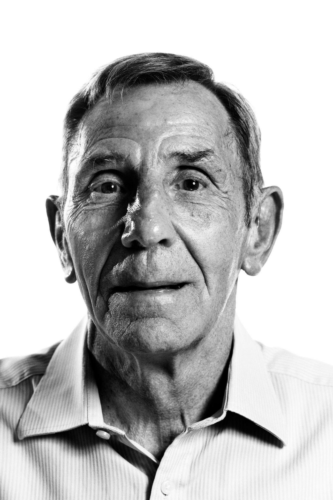 A black-and-white portrait of an elderly man looking directly into the camera. His hair is brushed to one side and he is wearing a light shirt.