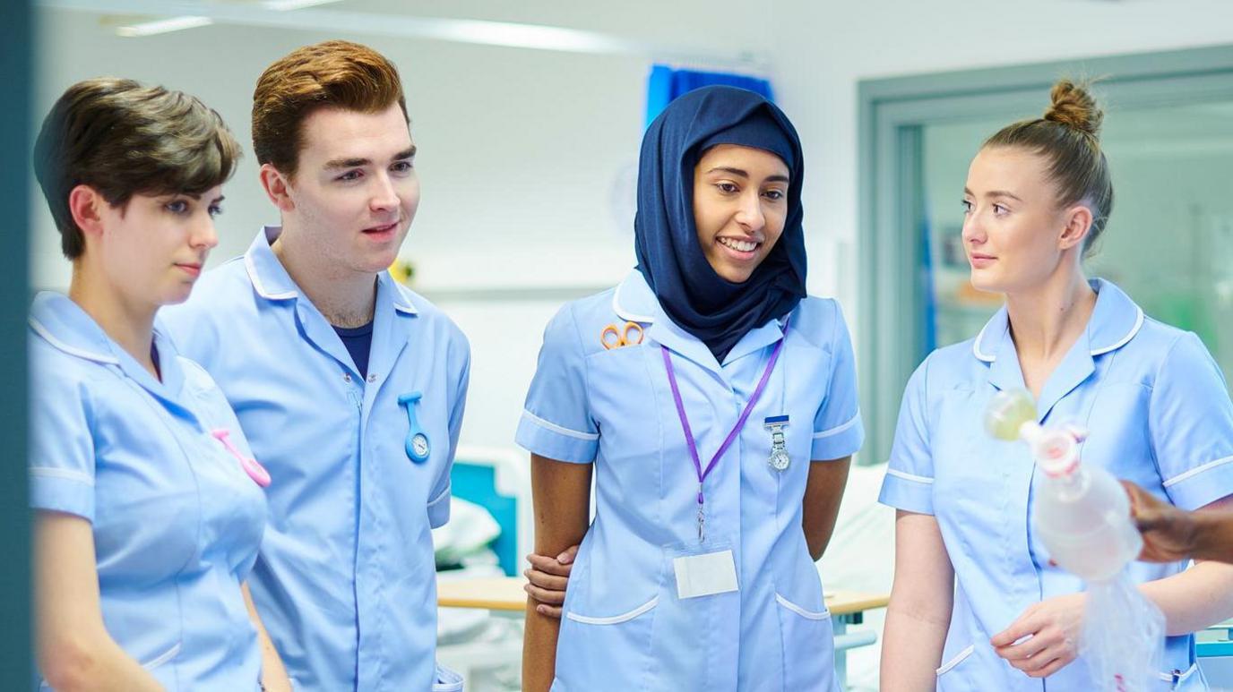 Four students are standing in a hospital ward wearing light blue uniforms, listening at another medic who is giving a demonstration. Each of the students has a watch pinned to their uniforms. The middle student also has a purple lanyard around her neck, and some scissors tucked into her top pocket.