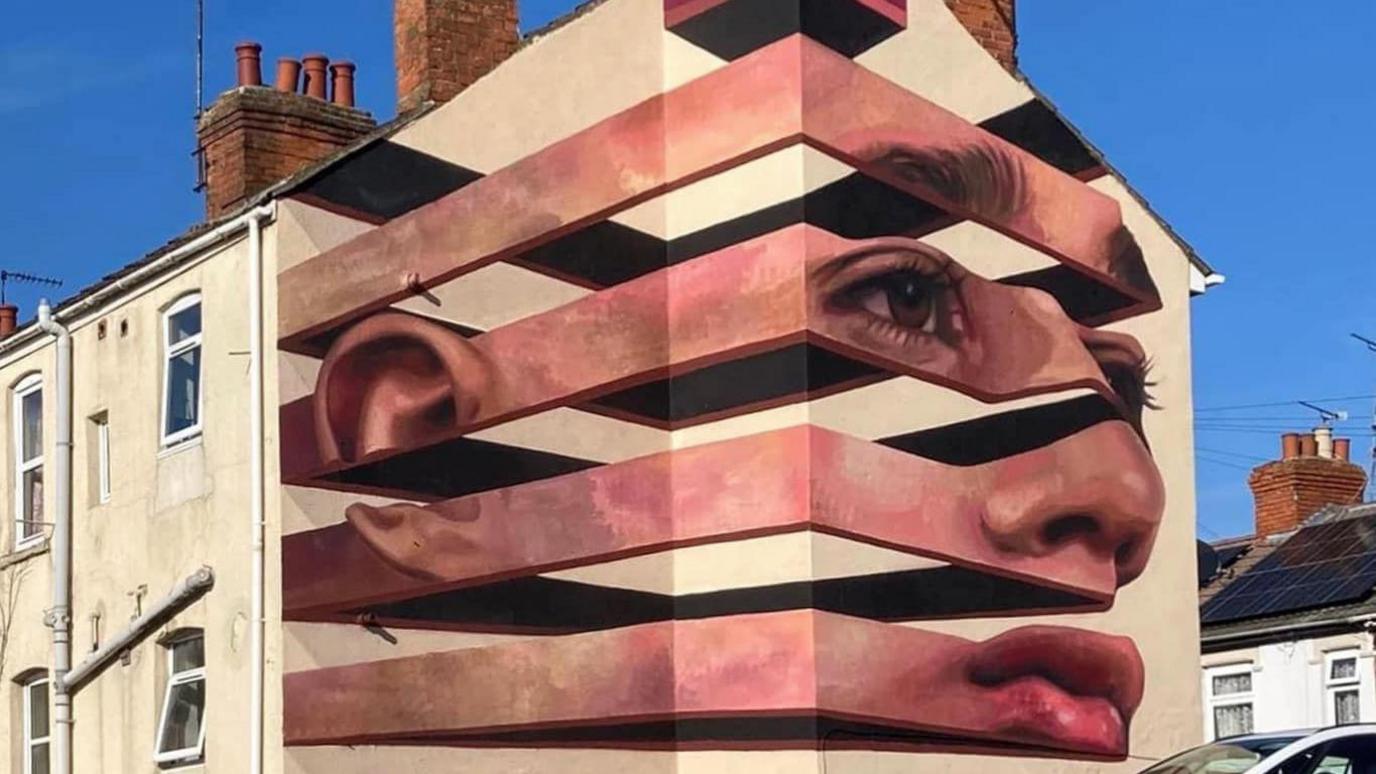 A mural on a cream coloured building which shows a woman's face in a cuboid shape which is slit into four parts with gaps of equal size in between