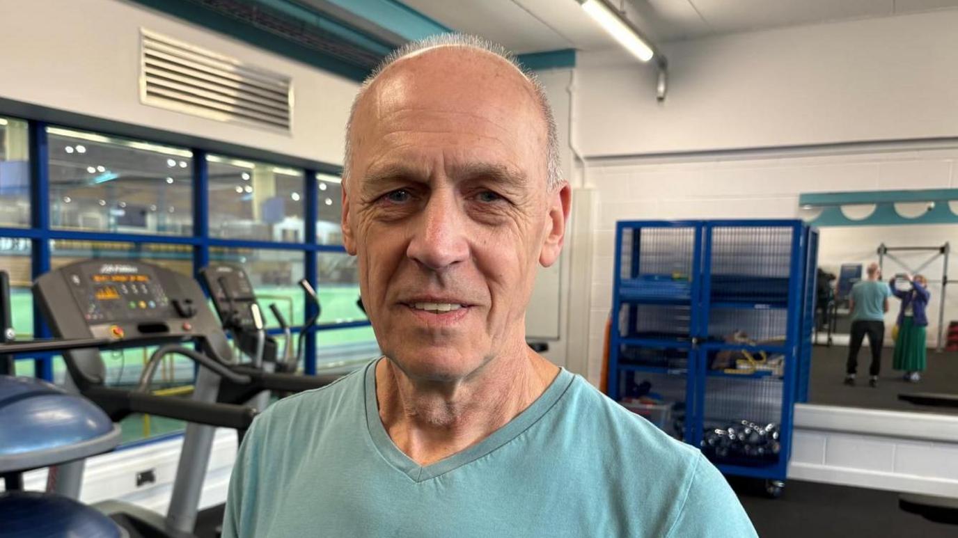 Anthony is stood in one of the gyms at Cardiff Met university, where he continues his physiotherapy since his cardiac arrest. Behind him are running machines and free weights. He is wearing a green t-shirt and looking at the camera. It is a head and shoulders shot of him 
