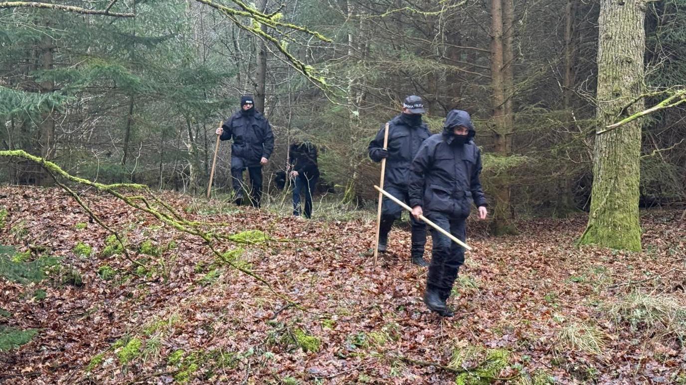 Four police officers dressed in black walking through a woodland. They're holding long wooden sticks.