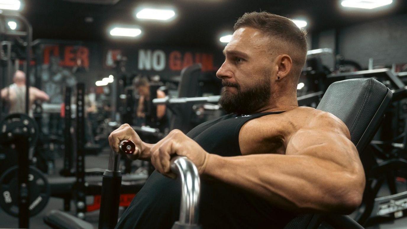 Brendan Pearl with his hands gripping handles on a machine where he is working his biceps.
