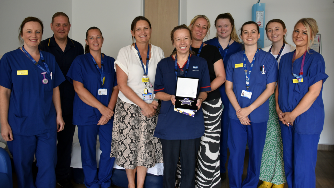 A group of midwifery staff in uniform, smiling to camera and surrounding Pip who is holding a certificate