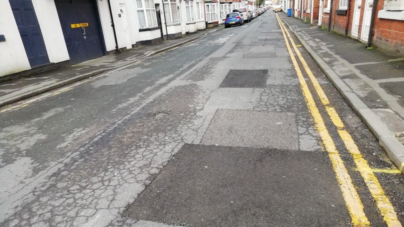 A patched up residential road. It has double yellow lines down both sides and terraced houses.