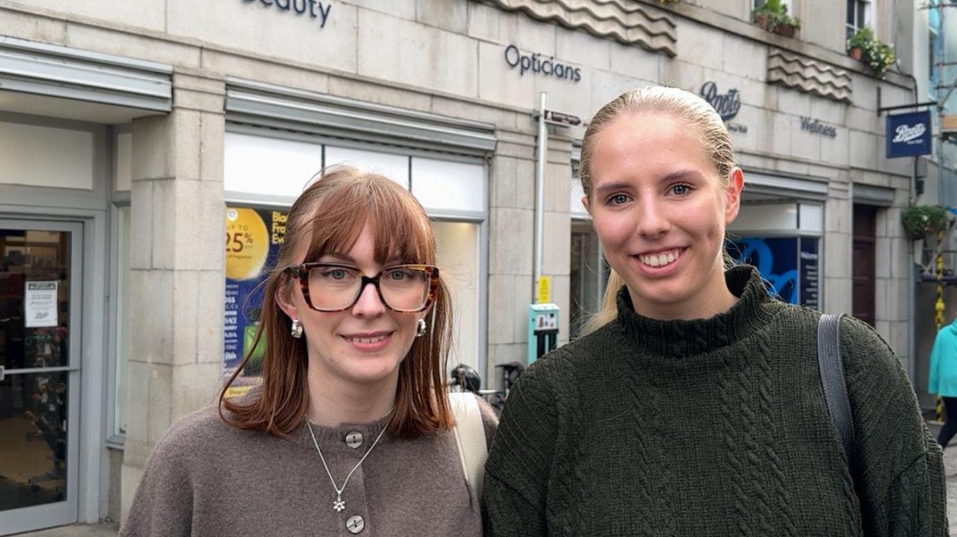Two woman in their 20s. One is slightly taller with a blonde pony tail and a green high neck jumper. The other has a brown long bob with a fringe and wears quite big square glasses