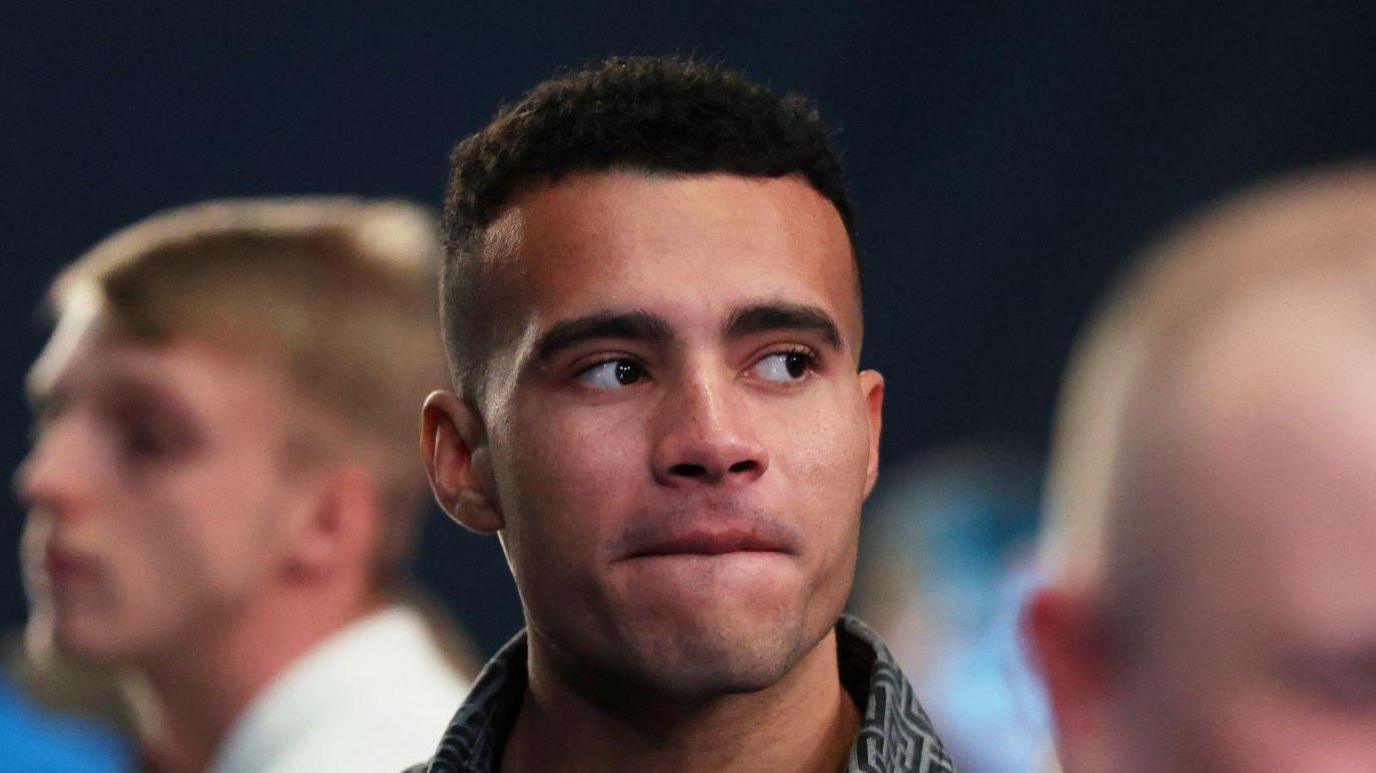 A young man with short black hair looking off to the right while stood in a crown of people.