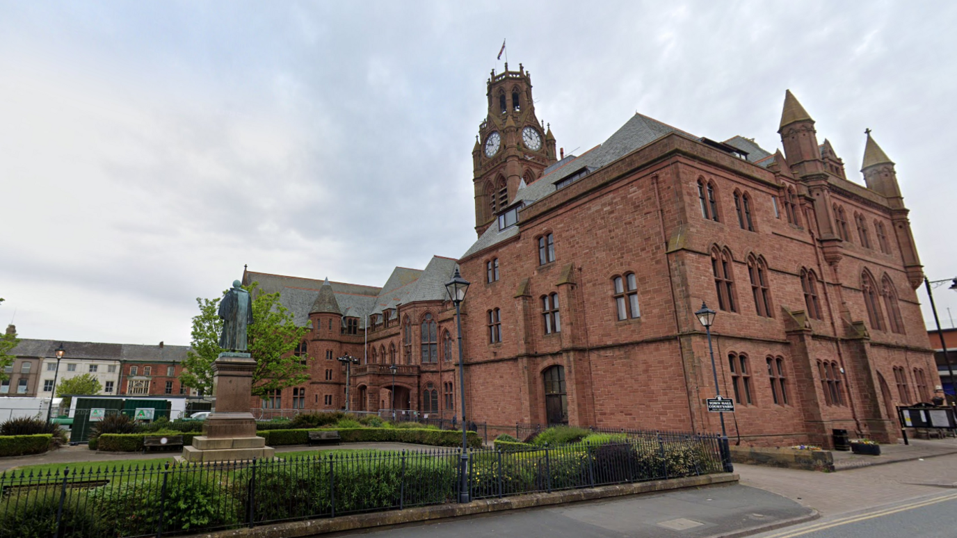 A general view of Barrow Town Hall. It is a large red-brick building with gothic features including turrets. In front of the building is a garden area with a statue.