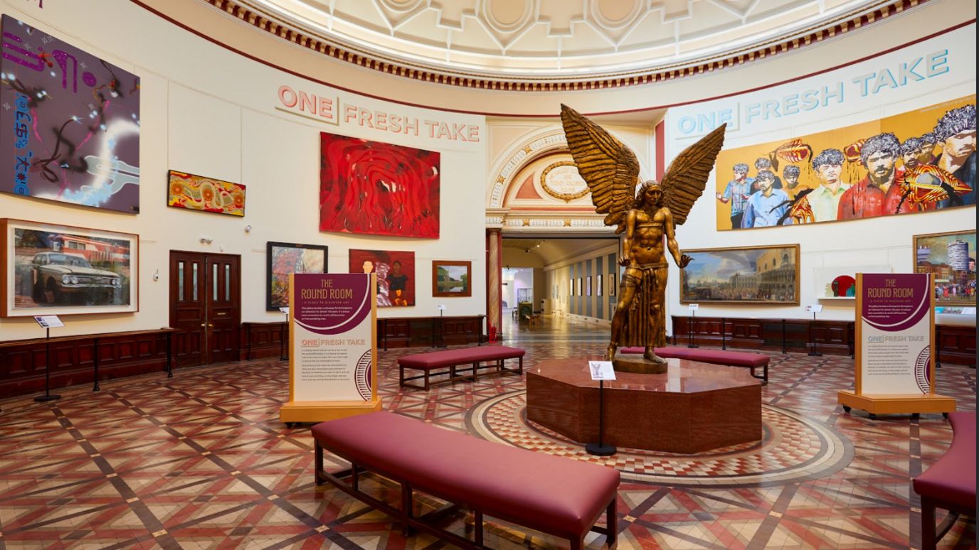 A circular room with a golden statue of an angel in the centre, it is surrounded by red benches and has two floor-standing signs either side of it. On the walls are multiple pieces of art, and at the top of the walls on both sides of the image are the words "One Fresh Take"