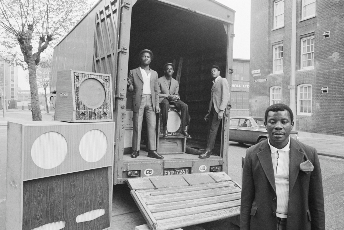 Black and white photograph of three men standing in the back of a truck parked next to a sound system. A man is also stood in front of the truck.