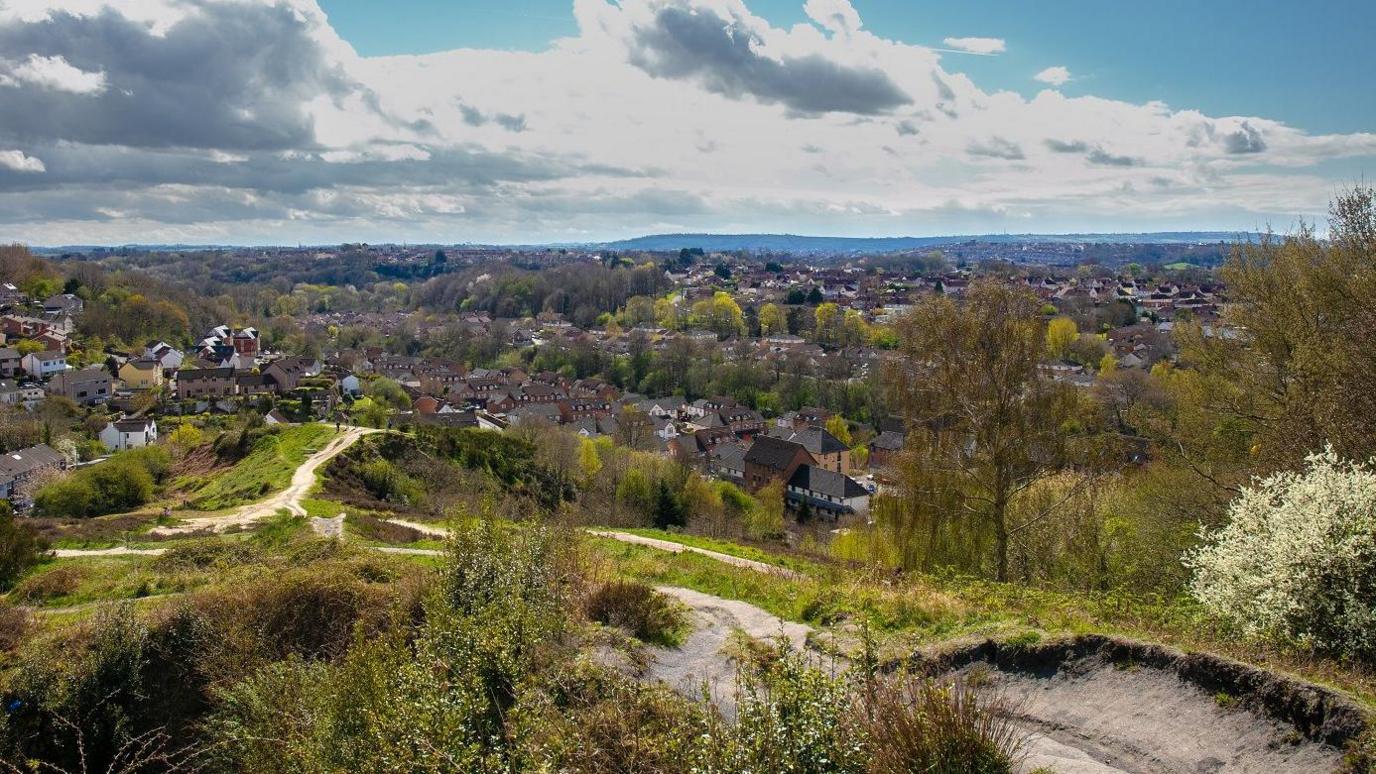 Panoramic view of a suburban area with houses surrounded by trees and greenery. Dirt paths wind through grassy and bushy terrain in the foreground. The background features forested areas and distant hills under a partly cloudy sky.
